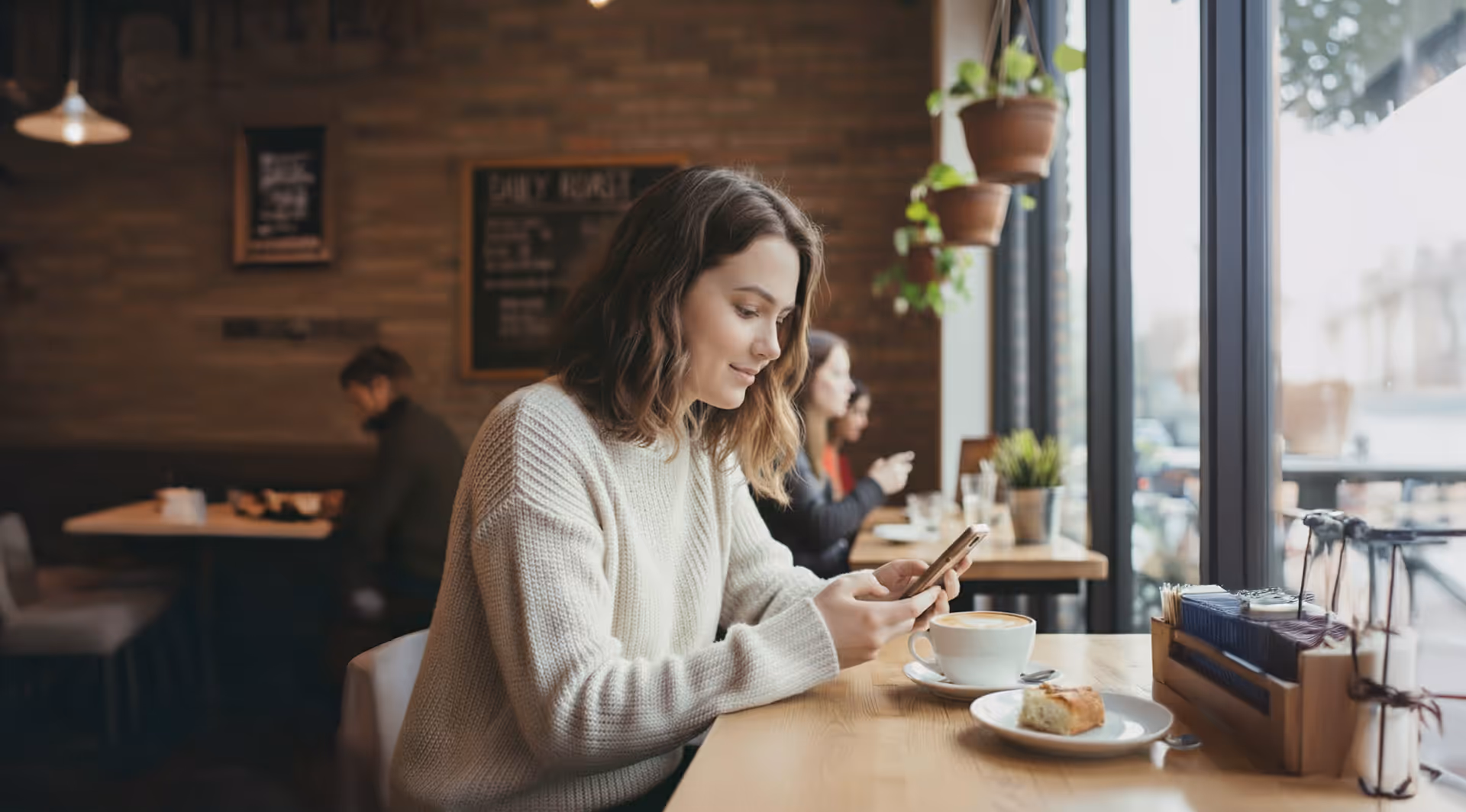 Woman in a white sweater sitting at a cafe table near a window, looking at her smartphone with a cup of coffee and a pastry in front of her.